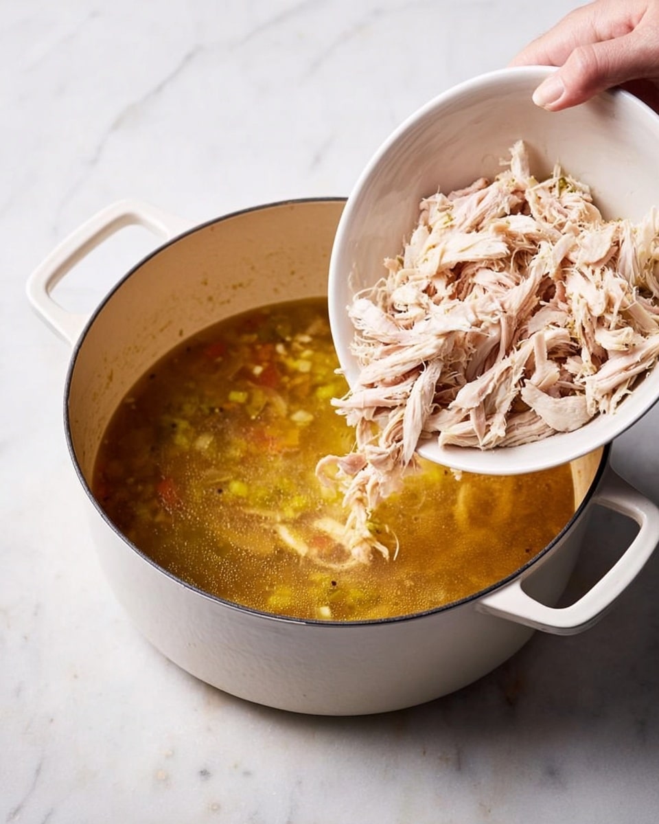 A white pot with two handles sits on a white marbled surface, filled three-quarters with golden brown broth containing small pieces of light yellow and green vegetables and spices. Above the pot, a woman's hand holds a white bowl tilted to pour a generous amount of shredded pale pink and white cooked chicken into the broth, with some falling into the liquid. The texture of the shredded chicken is soft and fibrous. Photo taken with an iphone --ar 4:5 --v 7