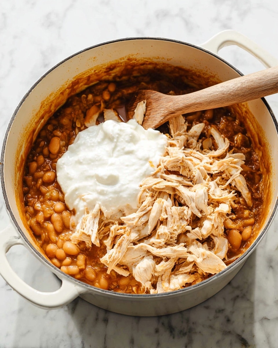 Inside a white pot, there is a layered dish with a thick base layer of brownish-orange beans in sauce, topped with shredded light beige chicken pieces scattered unevenly, and a fluffy white creamy layer placed right in the center on top. A wooden spatula rests inside the pot, partially submerged in the mixture, and the pot sits on a white marbled surface. photo taken with an iphone --ar 4:5 --v 7