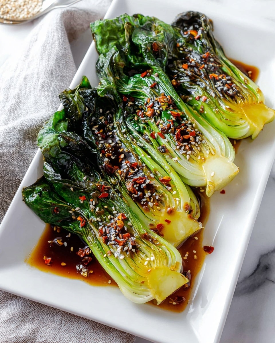 The dish shows four baby bok choy pieces, cooked and shiny with a dark soy sauce glaze, placed side by side on a white rectangular plate. The bok choy leaves are dark green, slightly wilted, and glossy while the stems are light green to pale yellow with a soft texture and slight browning from cooking. The sauce pools at the bottom of the plate, with small bits of red chili and sesame seeds sprinkled over the top and around the plate edges. The plate sits on a white marbled surface with a light gray cloth in the background. Photo taken with an iphone --ar 4:5 --v 7