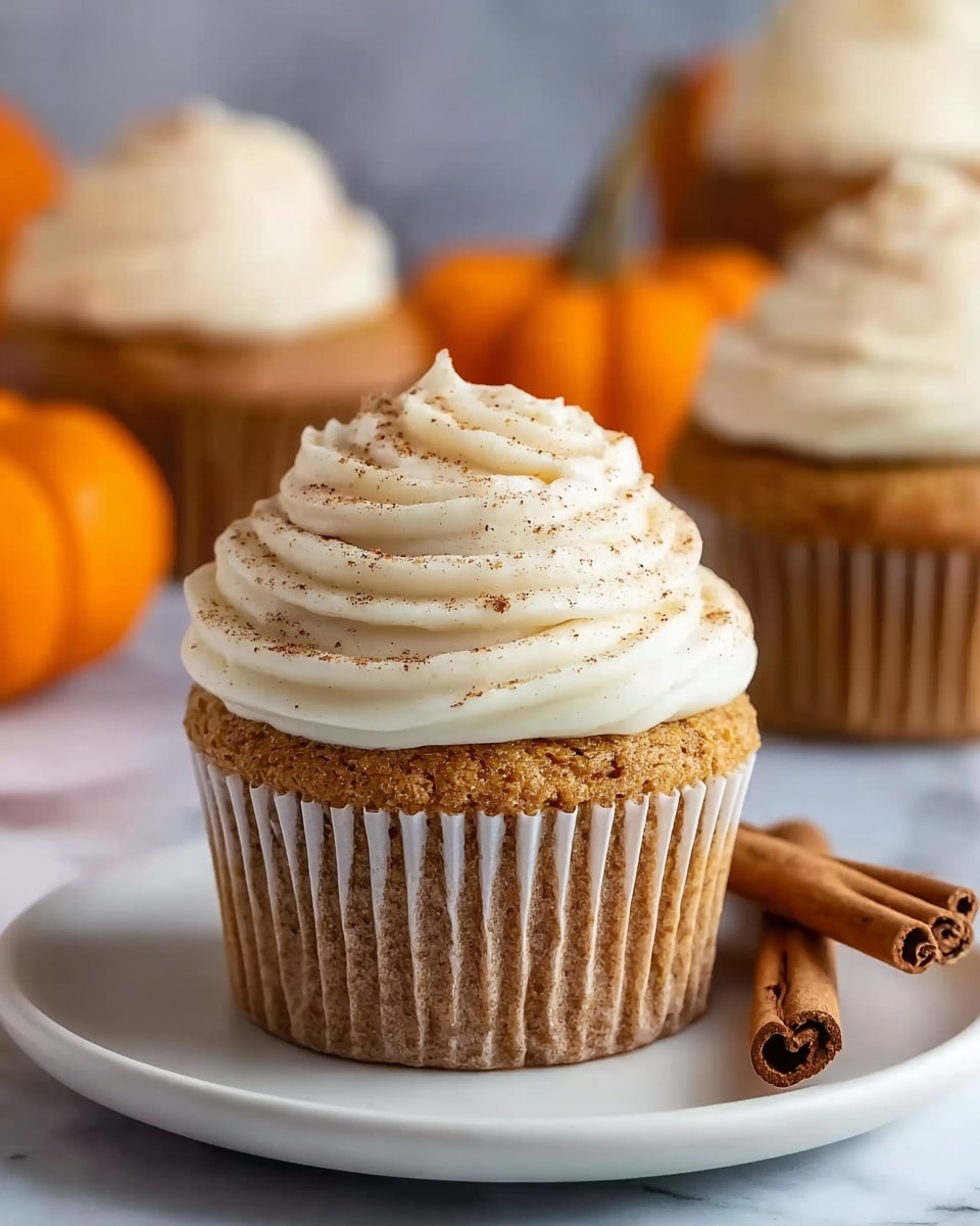 A single cupcake sits centered on a white plate with a white marbled texture background. The cupcake has one thick base layer of golden brown cake with a slightly rough texture and lined white paper wrapping. On top, there is a tall swirl of creamy white frosting, with smooth and thick texture spiraled neatly from the base to a small peak at the center. Light brown spice is lightly sprinkled over the frosting for decoration. Next to the cupcake on the plate are two cinnamon sticks placed diagonally. In the background, out of focus, more cupcakes with similar frosting and orange small pumpkins add a cozy, warm atmosphere. Photo taken with an iphone --ar 4:5 --v 7