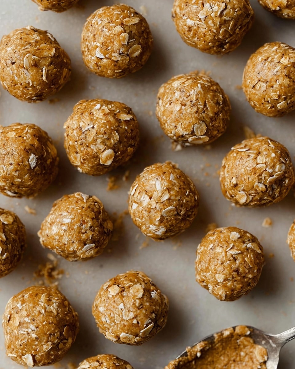 The image shows many small, round balls made of a light brown mixture with visible oats, giving them a rough, textured surface. They are arranged closely together on a flat white marbled background, filling the frame without any gaps. The balls appear firm and dense, with some slight cracks and oats sticking out, hinting at a chewy texture inside. In the bottom right corner, a bit of a silver spoon with a similar mixture is visible. The lighting is soft, highlighting the texture and warm color of the balls. Photo taken with an iphone --ar 4:5 --v 7
