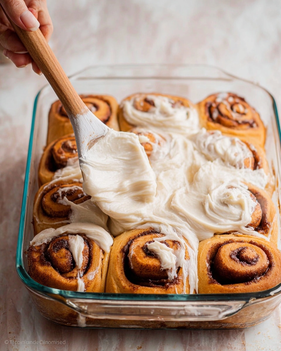 The image shows nine cinnamon rolls in a clear rectangular glass baking dish, arranged in three rows of three. The rolls are golden brown with dark cinnamon swirls visible inside each roll. A thick layer of creamy white frosting is being spread over the rolls with a wooden spoon held by a woman's hand with a white handle on the spoon. The spoon glides over the rolls, leaving a smooth and slightly textured frosting surface, partially covering most of the rolls. The background is a white marbled texture. photo taken with an iphone --ar 4:5 --v 7