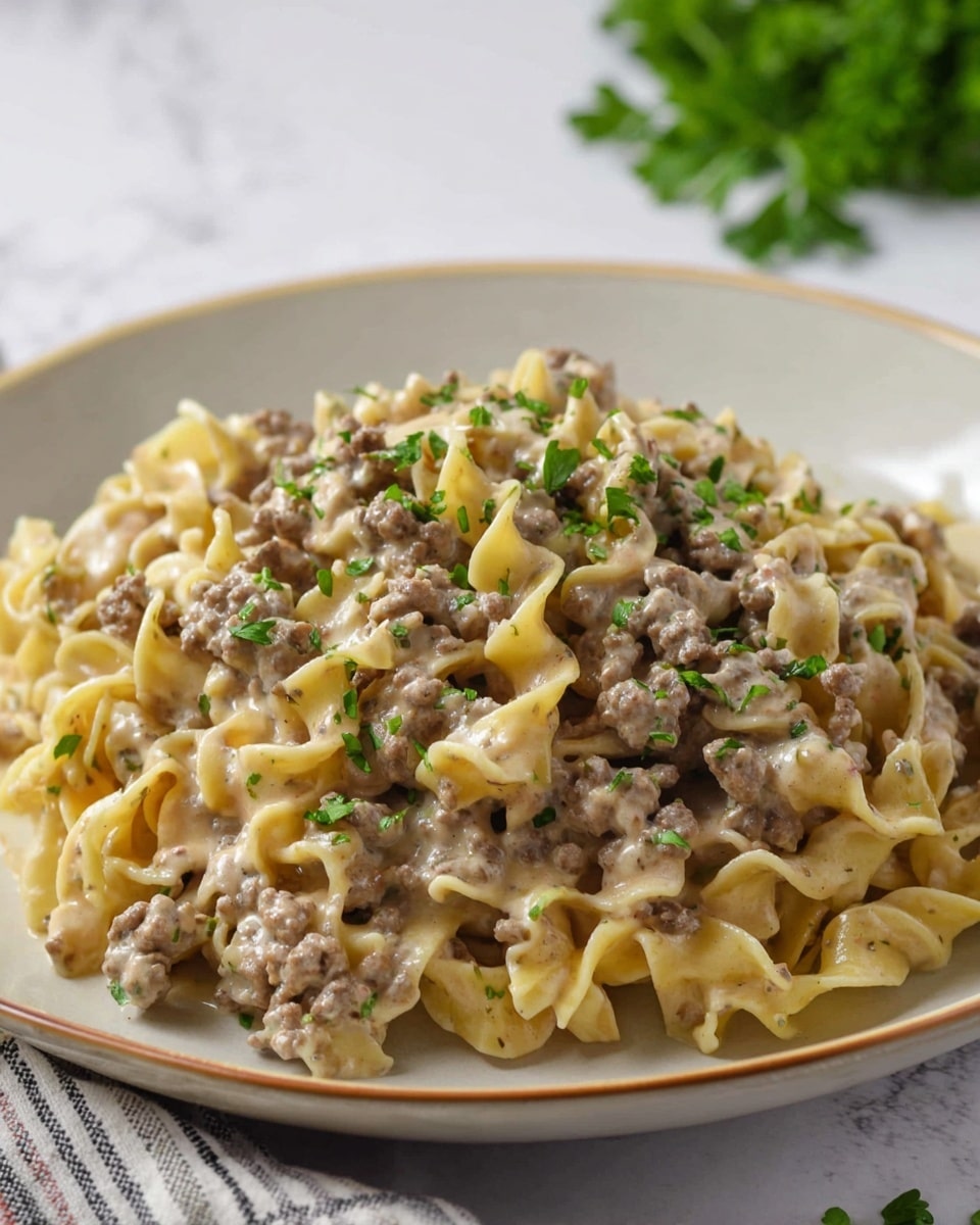 The dish shows a pile of twisted egg noodles in creamy light brown sauce mixed with cooked ground meat that appears soft and crumbly, evenly spread throughout. Bright green chopped herbs are sprinkled on top, adding a fresh color contrast. The food is served in a round white plate with a thin beige rim, placed on a white marbled surface. In the background, some green herb leaves are scattered softly out of focus. Photo taken with an iphone --ar 4:5 --v 7