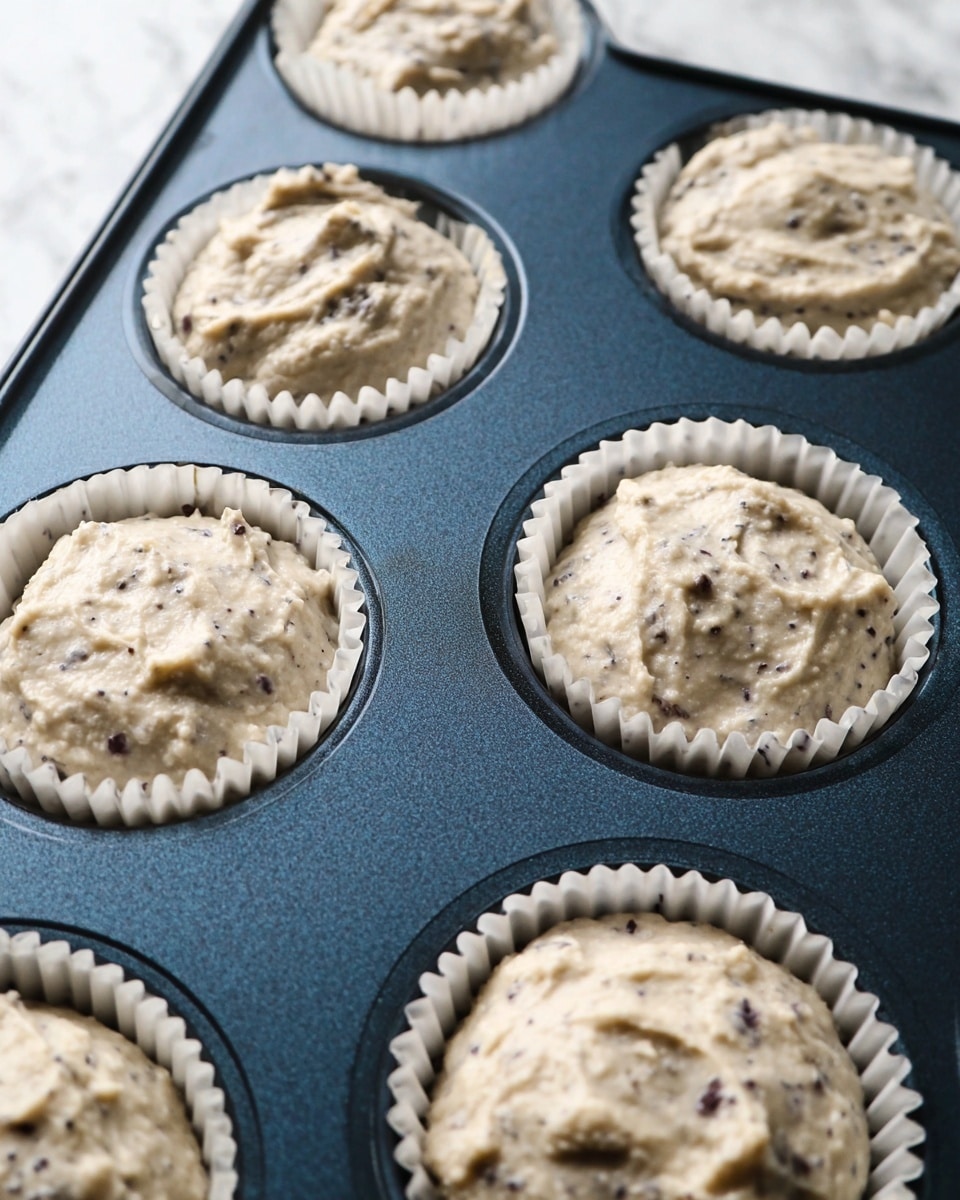 A close-up view of six muffin cups filled with thick, light beige batter with dark specks mixed throughout, sitting inside a dark blue metal muffin tray. Each cup is lined with white paper liners that contrast against the tray. The batter looks soft and slightly lumpy, filling each cup unevenly with some smooth and some rough patches. The background is a white marbled texture. photo taken with an iphone --ar 4:5 --v 7