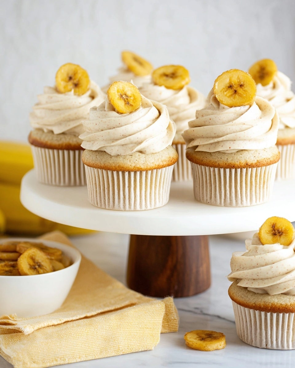 The image shows a white cupcake stand holding six cupcakes, each with three thick swirls of light beige frosting that has tiny specks visible in it. On top of each cupcake sits a crisp yellow banana chip, placed slightly tilted. The cupcakes have light golden-brown bases wrapped in white cupcake liners with a ridged texture. The stand has a round white top and a wooden brown base. Below the stand is a folded yellow cloth napkin with a couple of banana chips scattered on it. In the background, there is a white bowl filled with more banana chips, all set against a white marbled surface. photo taken with an iphone --ar 4:5 --v 7