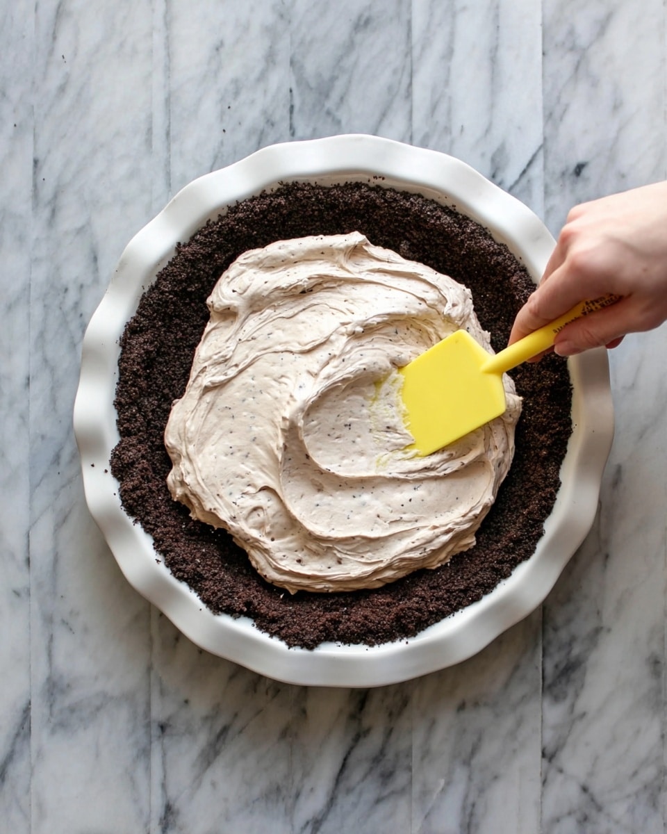 A white pie dish holds a dark crumbly crust layer around the edges and bottom, with a thick, light brown creamy filling being spread gently by a woman's hand holding a yellow spatula. The creamy filling has a smooth, fluffy texture with some small dark specks mixed in, sitting centrally on top of the crust. The dish is placed on a white marbled surface. Photo taken with an iphone --ar 4:5 --v 7