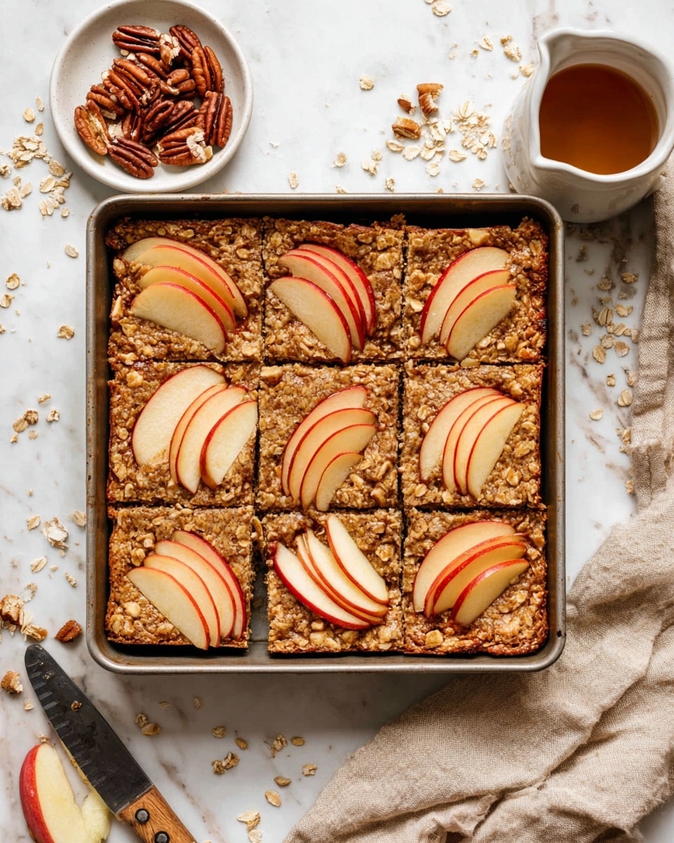 A square baking pan with six rectangular oat bars inside, each bar topped with thin apple slices arranged in a fan shape; the oat bars have a rough, crumbly texture with visible chunks of apple mixed in, colored light brown with hints of red from the apples. The pan rests on a white marbled surface scattered with pieces of oats and pecans, next to a small white plate filled with broken pecan pieces, a small white bowl with syrup, and a beige cloth with a knife. Photo taken with an iphone --ar 4:5 --v 7