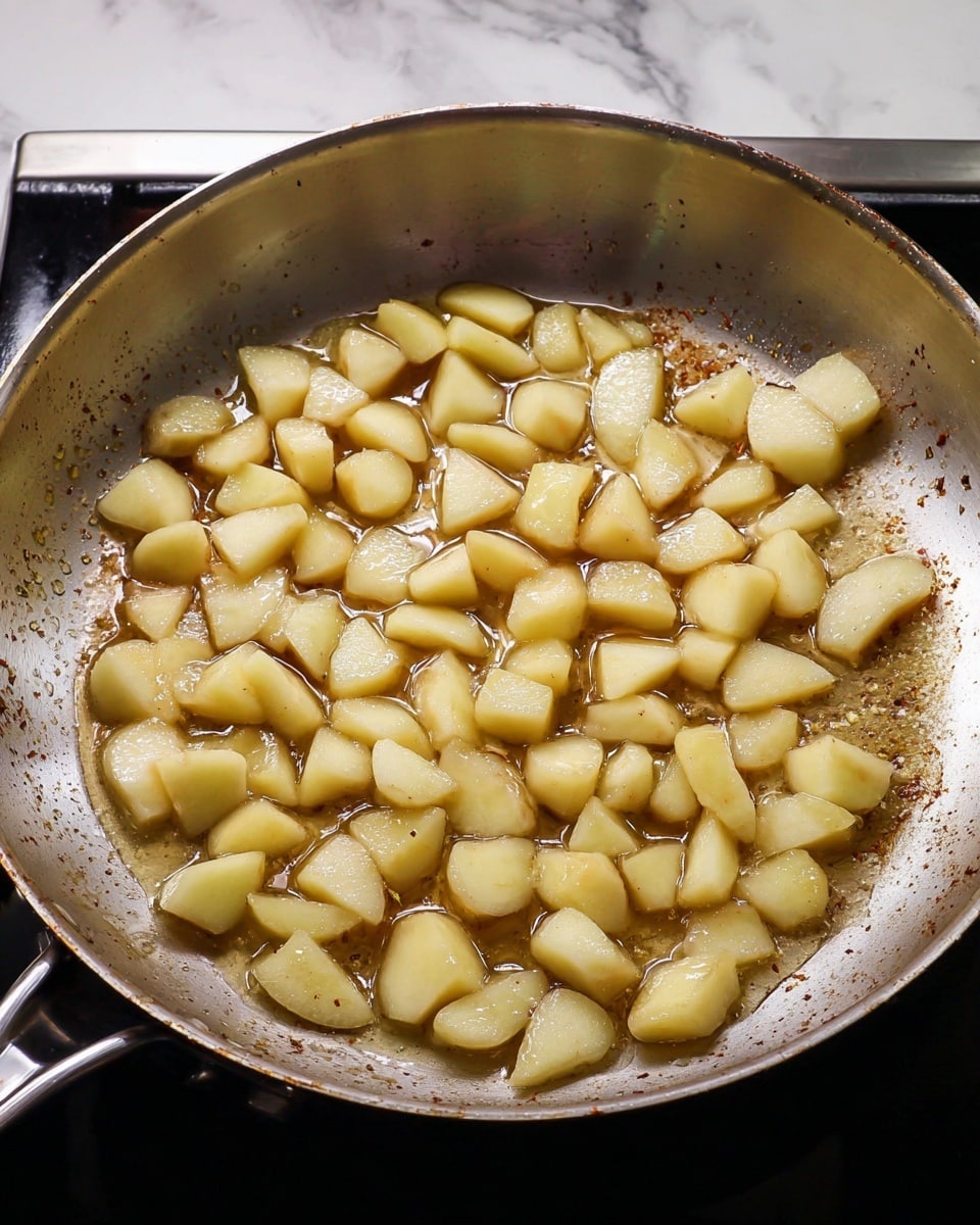 A metal frying pan filled with a single layer of small, soft, pale yellow apple pieces cooking in a thin light brown syrupy liquid. The apple pieces have smooth surfaces with slight browning on some edges, and the pan’s inside shows some cooked-on residue. The pan sits on a black stove, but the background is changed to a white marbled texture. photo taken with an iphone --ar 4:5 --v 7