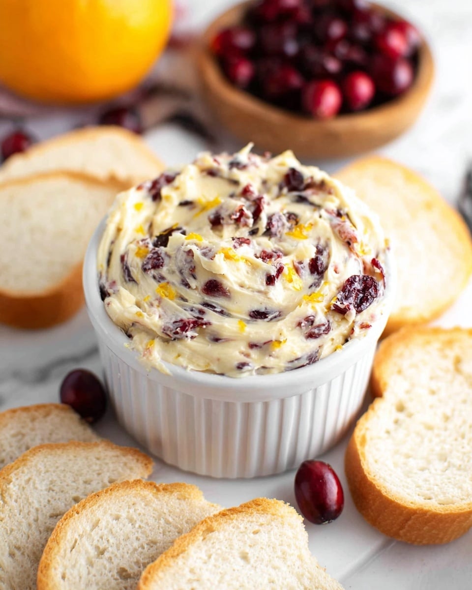 A white ramekin is filled with creamy butter mixed with small pieces of dark red cranberries and hints of orange zest, giving it a textured look with swirls and specks throughout. Surrounding the ramekin are several slices of light, soft white bread arranged casually on a white marbled surface. In the background, half an orange and a wooden bowl with more cranberries add color and context to the scene. The overall setting is bright and clean with a fresh, inviting appearance. photo taken with an iphone --ar 4:5 --v 7