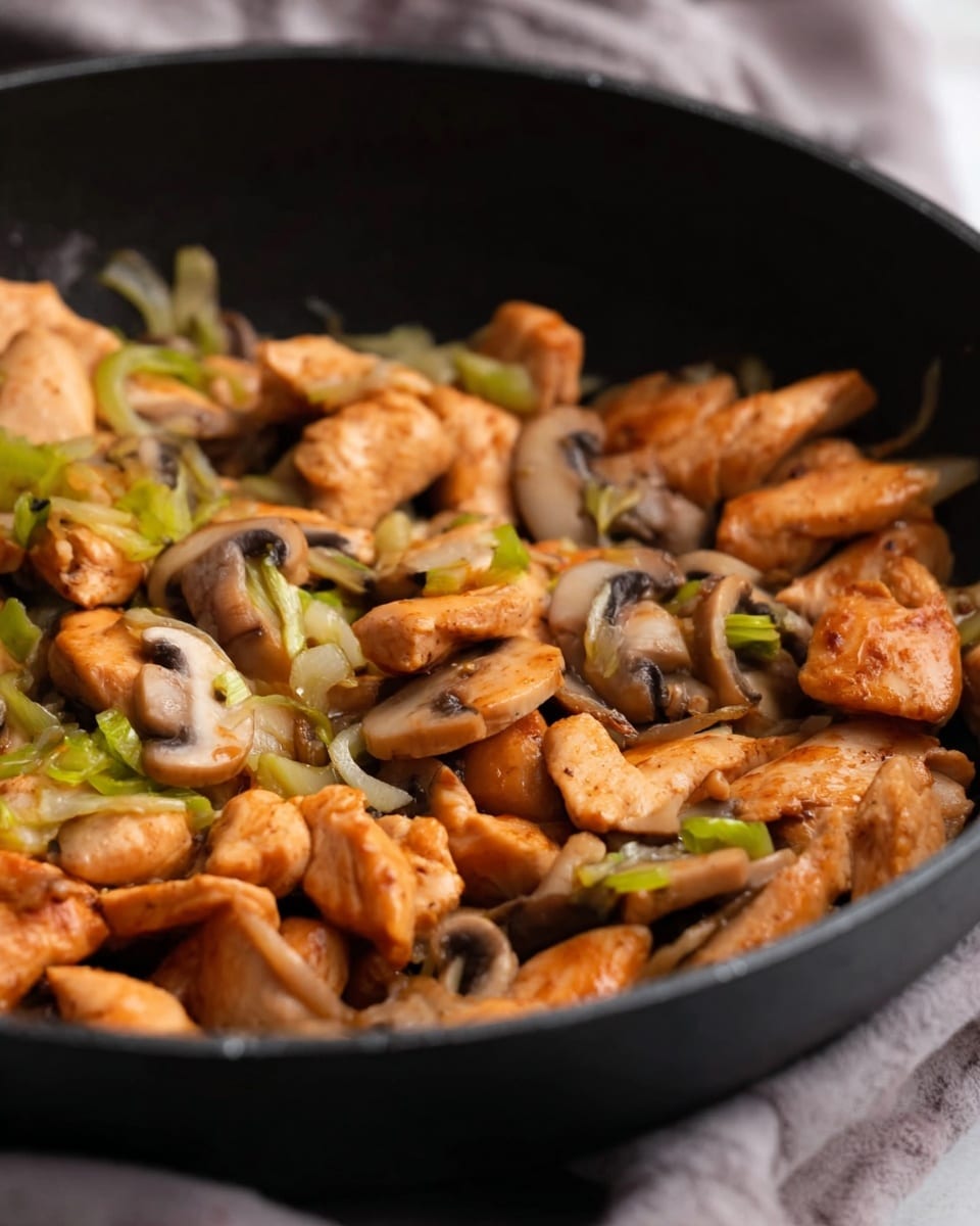 Inside a black pan, small pieces of cooked chicken are mixed with sliced mushrooms and thin strips of light green vegetables. The chicken chunks are golden brown with a slightly crispy texture, while the mushrooms have a soft, smooth look with light and dark brown shades. The green vegetable pieces add a fresh touch that contrasts with the warm colors of the chicken and mushrooms. The background shows a soft, blurred cloth making the food the main focus. Photo taken with an iphone --ar 4:5 --v 7