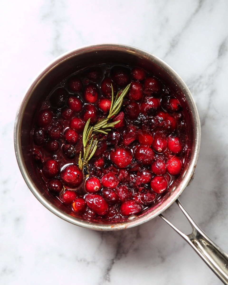 A silver cooking pot viewed from above sits on a white marbled surface, filled with cooked red cranberries and a sprig of rosemary. The cranberries are soft, some darker, some bright red, mixed in a thick sauce that coats the bottom of the pot. The pot handle extends to the right side, showing its smooth metallic texture. photo taken with an iphone --ar 4:5 --v 7
