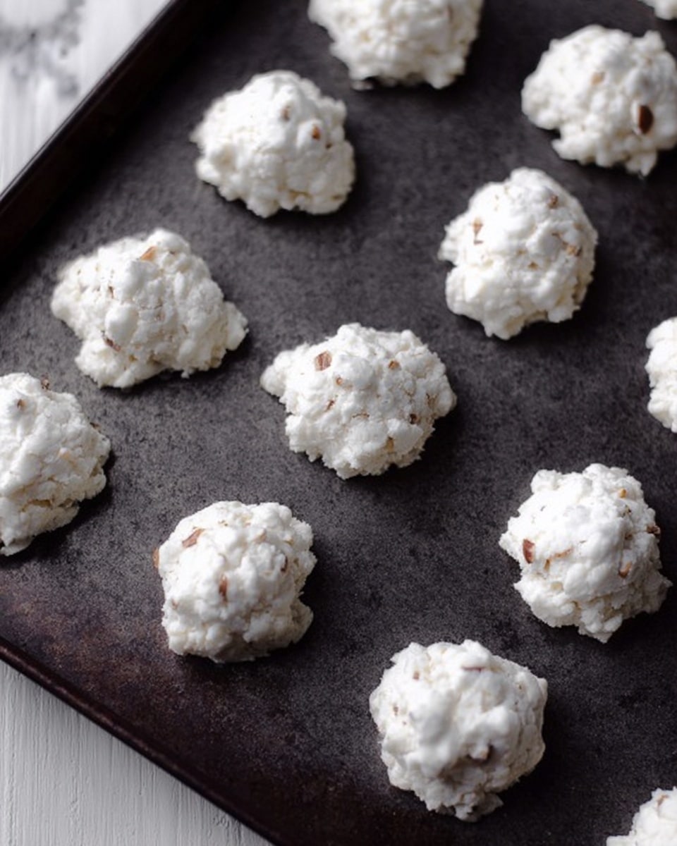 The image shows a dark baking tray filled with small clusters of white, crumbly cookie-like mounds scattered evenly. Each mound has a rough, uneven surface with little bumpy textures, showing small nut pieces inside the white mixture. The dark tray contrasts with the bright white clusters. The background is a white marbled surface. photo taken with an iphone --ar 4:5 --v 7