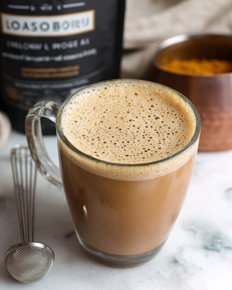 A clear glass mug filled with a light brown frothy drink with many small bubbles on the top layer, sitting on a white marbled surface. Behind the mug, there is a partially visible black and white package with text, and a blurred round container with a golden brown finish. A small metal whisk tool lies on the white marbled surface in front of the mug. The scene is softly lit, emphasizing the creamy texture of the drink. Photo taken with an iphone --ar 4:5 --v 7