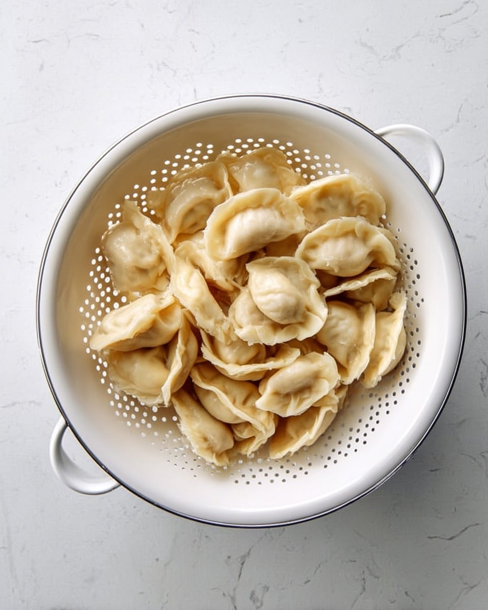 Top view of a white colander filled with cooked dumplings that are pale beige in color with smooth and slightly wrinkled texture. The dumplings vary in shape, some round with folds on top, others crescent-shaped with curved edges. The colander has holes around its sides and is placed on a white marbled surface. Photo taken with an iphone --ar 4:5 --v 7