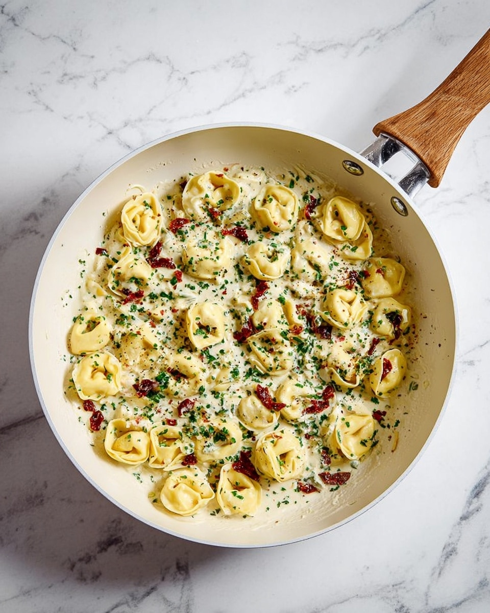 A white pan with a wooden handle is filled with about two layers of plump, yellow tortellini pasta covered in a creamy, white sauce. Small bits of red sun-dried tomatoes and green herbs are scattered evenly across the top, adding pops of color and texture. The pasta looks soft and slightly shiny from the sauce, with some herbs sprinkled more densely near the edges. The pan sits on a white marbled surface, showing a clean and bright kitchen setting. photo taken with an iphone --ar 4:5 --v 7