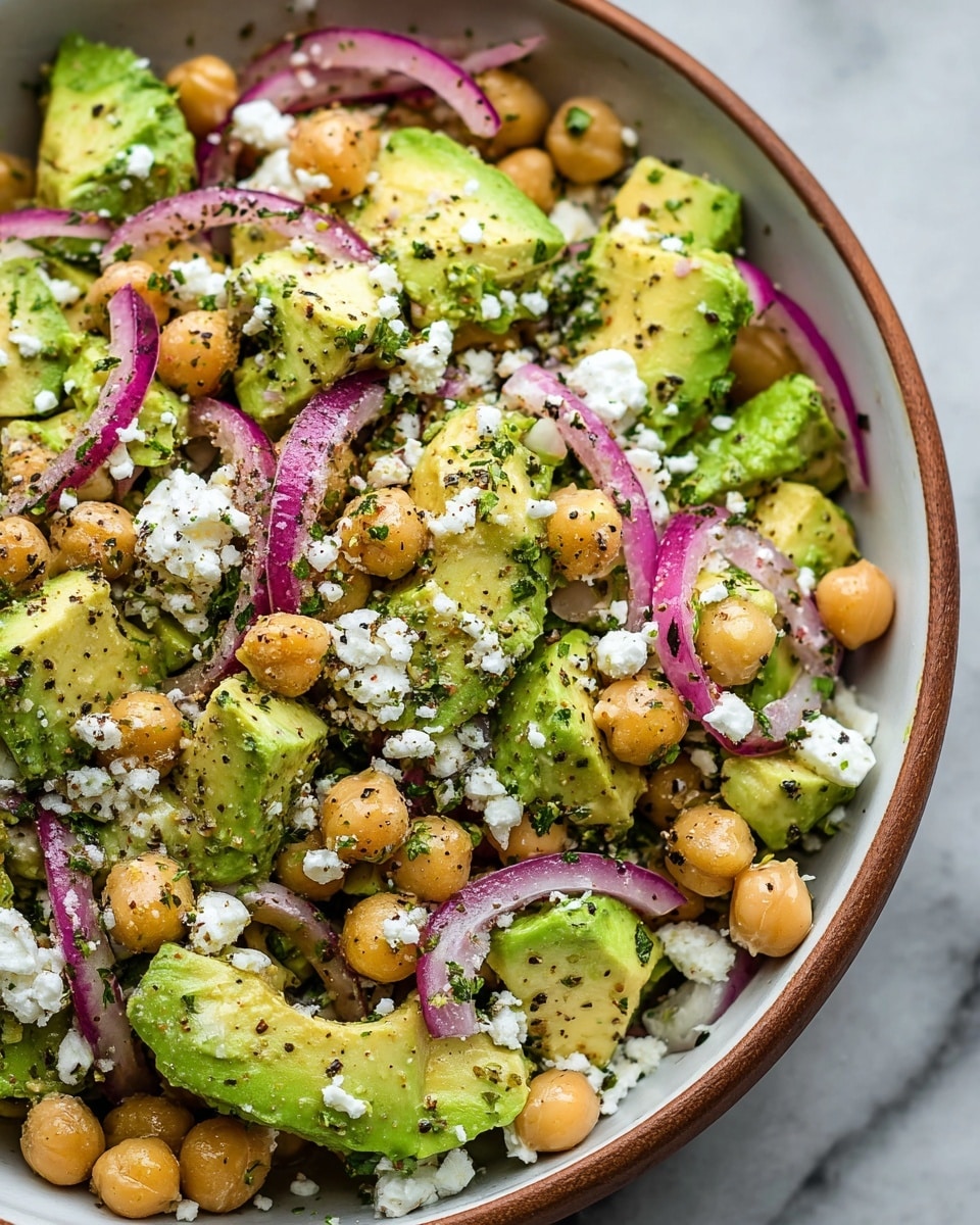 A close-up view of a creamy avocado and chickpea salad placed inside a white bowl with a wooden rim. The salad has large bright green avocado chunks scattered evenly throughout, mixed with whole light tan chickpeas. Thin slices of purple-red onion are spread across the salad, adding a contrast in color. White crumbled cheese is sprinkled on top, mingled with finely chopped green herbs and black pepper bits, creating a fresh, textured look. The onion slices and herbs add a slight shine, and the mix looks vibrant and fresh. The background is a white marbled surface. Photo taken with an iphone --ar 4:5 --v 7