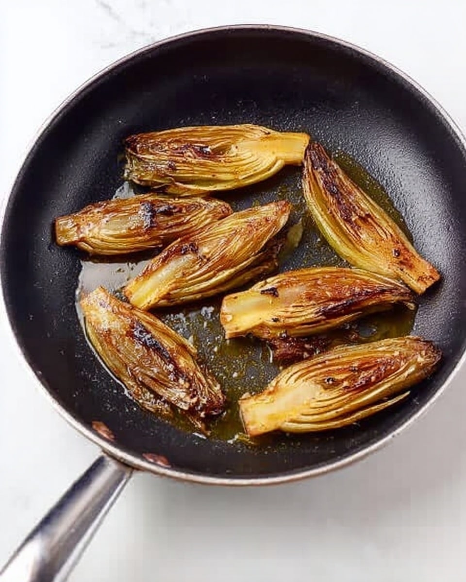 A black frying pan holds eight pieces of cooked endive, each cut lengthwise in half. The endives are golden brown and slightly charred on the edges, showing a caramelized texture. The pan's shiny metal handle points left, and the pan sits on a white marbled surface. The inside of the pan has some oil glistening under the endive pieces. photo taken with an iphone --ar 4:5 --v 7
