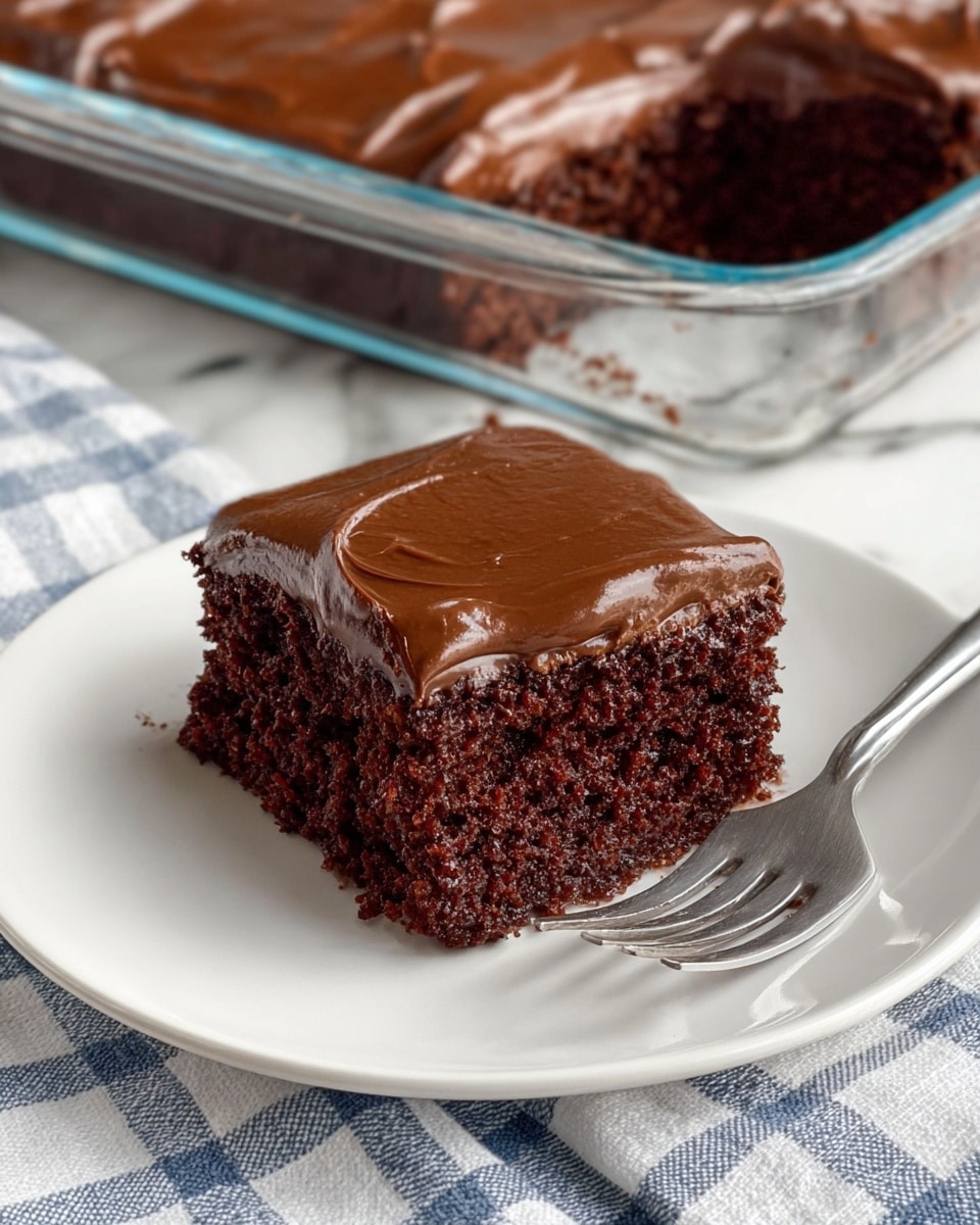 A single piece of chocolate cake is shown on a white plate with one layer of dark brown, moist cake topped with one thick, smooth layer of glossy chocolate frosting. The cake texture looks soft and slightly crumbly. Behind the plate, a silver fork rests on the plate edge, and in the background, a larger glass dish with clear sides holds more chocolate cake with the same frosting. The surface below the plate is a white marbled texture covered partially with a white and blue checkered cloth. Photo taken with an iphone --ar 4:5 --v 7