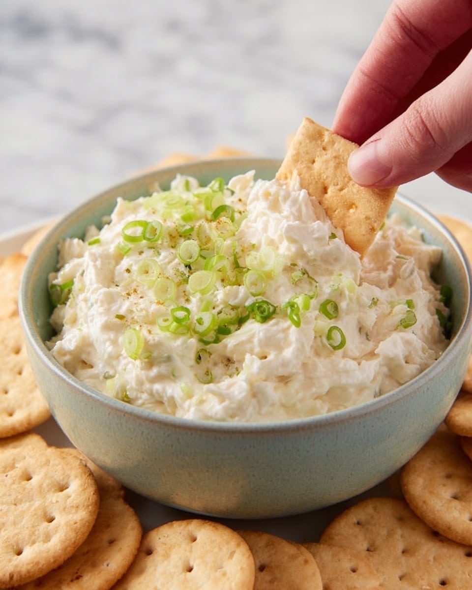 A round bowl with a smooth light grayish green color holds a creamy white dip with a soft texture. The dip is topped with small pieces of green onions scattered on the surface. The bowl sits on a large white tray filled with three types of crackers arranged in neat rows around the bowl: square light brown crackers with small holes, round pale beige crackers with a slightly rough texture, and square dark beige crackers with dark spots. The tray is placed on a white marbled surface with a soft cloth and a spoon nearby. Photo taken with an iphone --ar 4:5 --v 7