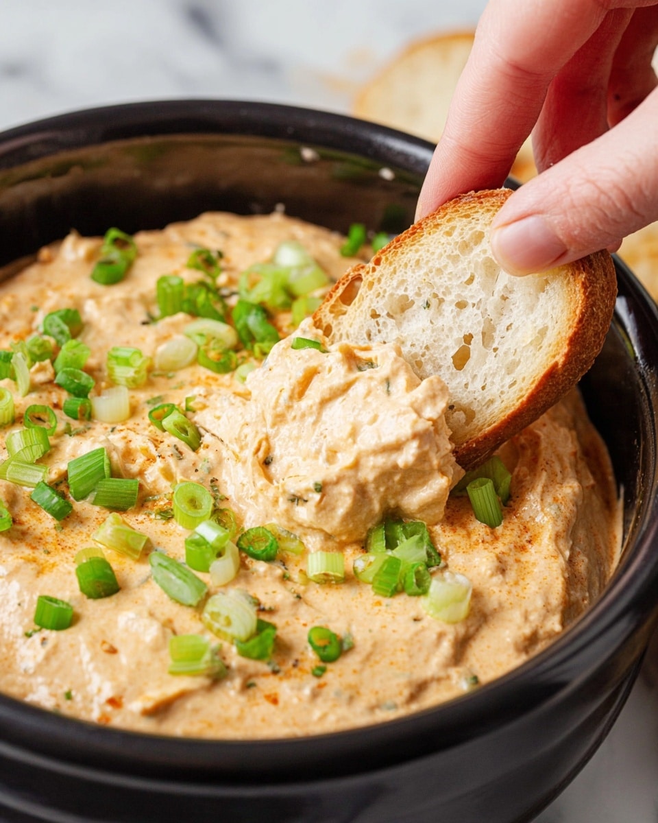 A close-up view of a black bowl filled with a thick, creamy dip that has a light orange color with visible small specks of darker seasoning throughout. The dip is topped with bright green, chopped scallions scattered evenly over the surface. A woman's hand is dipping a small piece of toasted, light golden brown bread with a porous texture into the creamy dip. The bowl is set on a white marbled surface. photo taken with an iphone --ar 4:5 --v 7