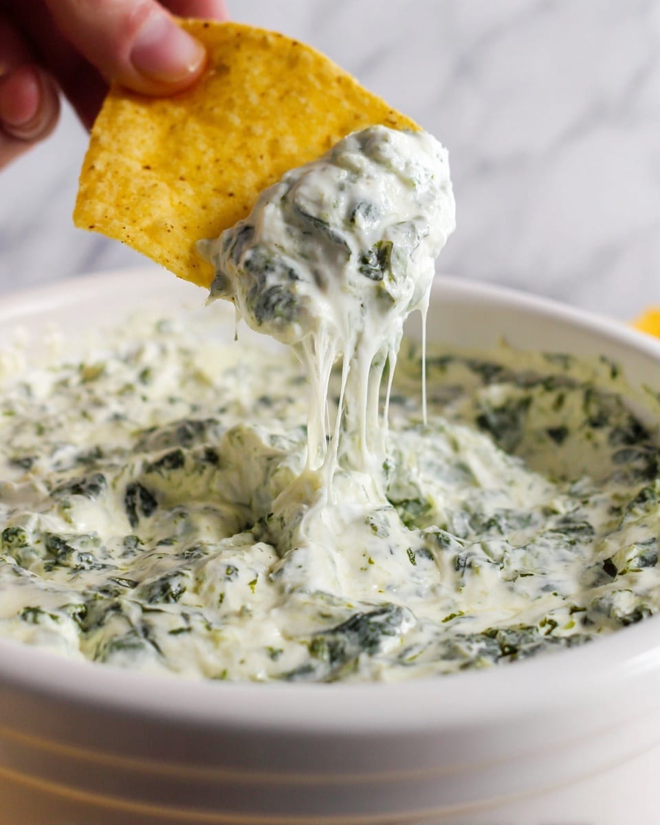 A close-up image shows a white bowl filled with a creamy spinach dip that has a thick, smooth texture with visible bits of green spinach spread evenly throughout. A yellow tortilla chip is dipped into the dip, lifting some of it with strands of melted cheese stretching from the bowl to the chip. A woman's hand holds the chip from the top left corner, adding a human touch. The background is simple with a white marbled surface that keeps the focus on the dip and chip. photo taken with an iphone --ar 4:5 --v 7