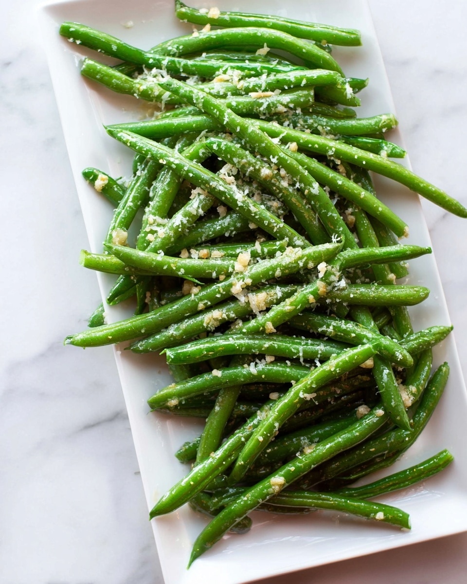A white rectangular plate filled with bright green cooked green beans arranged in a loose pile. The beans are coated lightly with a shiny glaze and sprinkled with small bits of minced garlic and fine grated cheese, which adds a pale yellow texture on top. The plate rests on a white marbled surface. The photo taken with an iphone --ar 4:5 --v 7