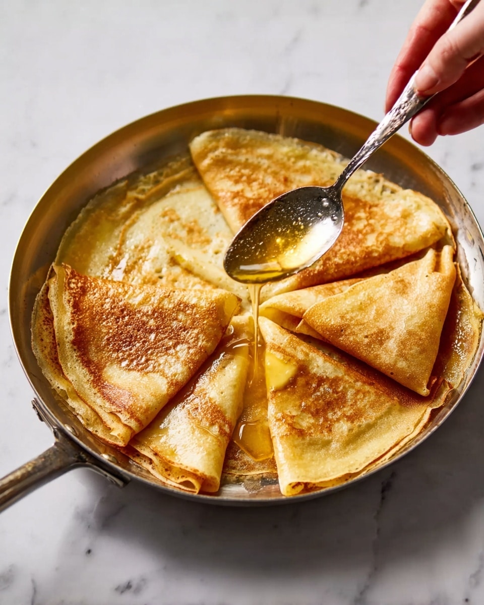 The image shows a white plate with a thin blue line on the edges, filled with many folded, golden, slightly shiny crepes soaked in a light brown sauce. The crepes are layered in a way that some are folded into triangles while others are folded into rectangles, and they cover the whole plate. Two old silver spoons are placed on the right side of the plate, resting partly on the crepes. The plate is on a light wooden table, next to a small white plate with some yellow sauce and crepes, a blue glass, and a white coffee cup with black coffee. The photo taken with an iphone --ar 4:5 --v 7