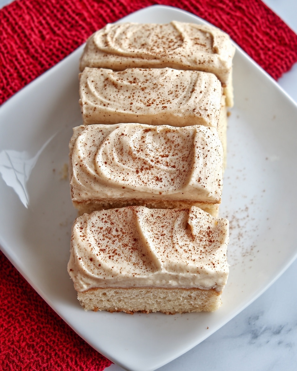The image shows four rectangular slices of a light beige cake arranged vertically on a white plate. Each slice is topped with a thick, creamy layer of off-white frosting that has a smooth, slightly textured surface with swirled patterns. The frosting on each slice is lightly sprinkled with a dusting of brown powder, evenly spread in small amounts. The plate is placed on a white marbled surface with a bright red textured cloth partially visible on the left side. The photo taken with an iphone --ar 4:5 --v 7