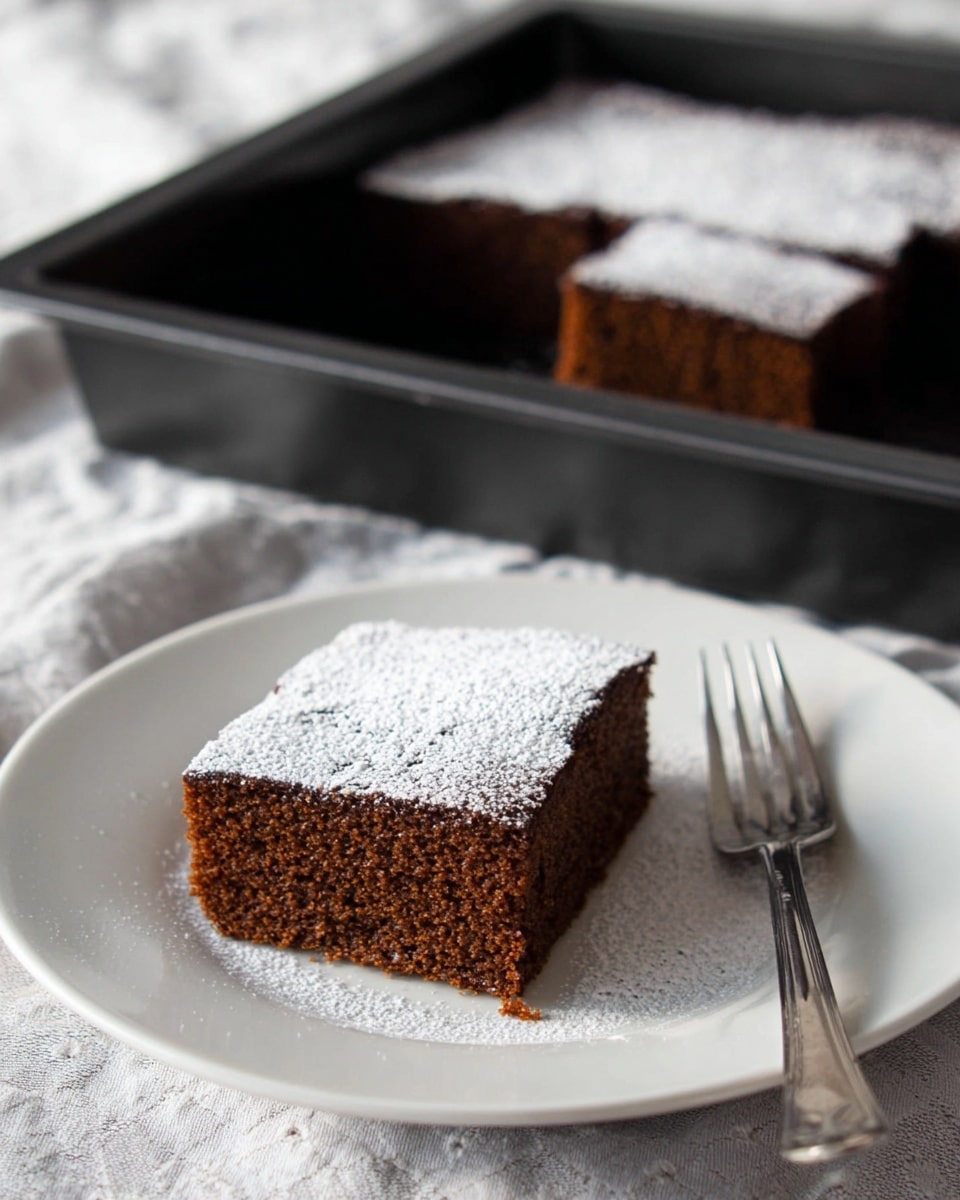 A single square piece of dark brown cake with a soft and moist texture is placed in the center of a white plate. The top of the cake is covered evenly with a thin layer of white powdered sugar. Next to the cake on the right side of the plate is a simple silver fork. In the background, there is a black baking tray holding the rest of the cake with a square piece already removed. The whole scene is set on a white marbled textured cloth surface. photo taken with an iphone --ar 4:5 --v 7