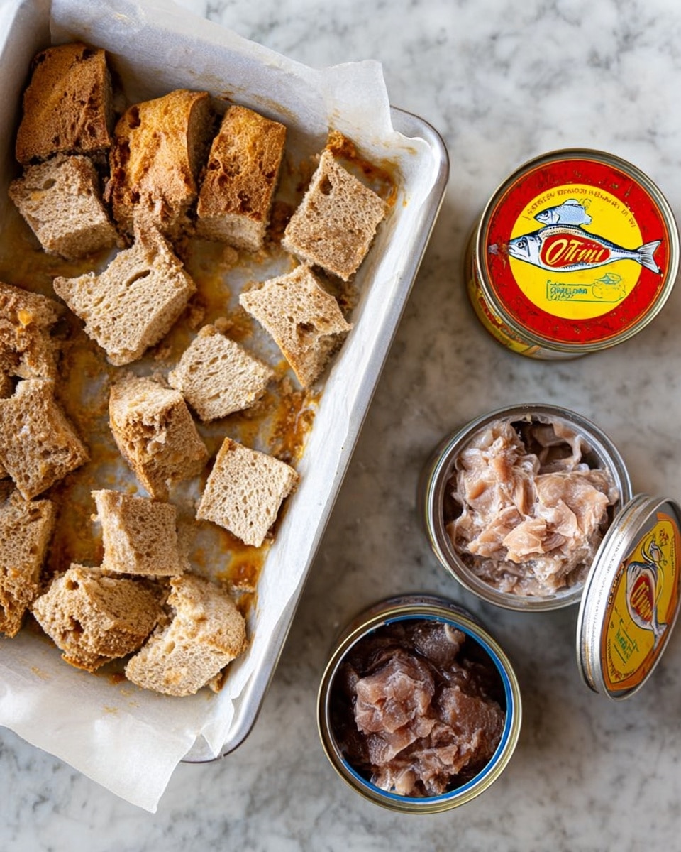 A white baking tray is filled with torn pieces of golden brown bread that have a rough texture and a slightly crispy top layer, placed on parchment paper. Next to the tray on a white marbled surface are three open small cans of fish with metallic lids peeled back. One can has dark brown fish packed tightly, the second has light pinkish fish flakes layered inside, and the third has a bright yellow and red label with a small bright fish illustration on top of the closed lid. The scene shows a mix of rough bread and shiny metal containers on a white marbled background. photo taken with an iphone --ar 4:5 --v 7