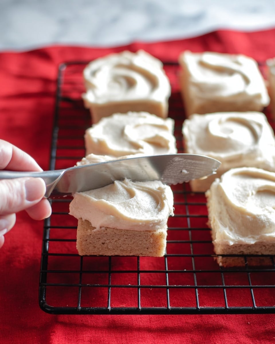 A woman's hand is holding a small, square piece of light brown dough with a slightly rough texture. On top of the dough, a thick layer of creamy, white frosting is being spread smoothly with a metal knife. In the background, more square dough pieces with uneven swirls of the same white frosting are placed on a black cooling rack over a bright red cloth. The surface beneath is a white marbled texture. photo taken with an iphone --ar 4:5 --v 7