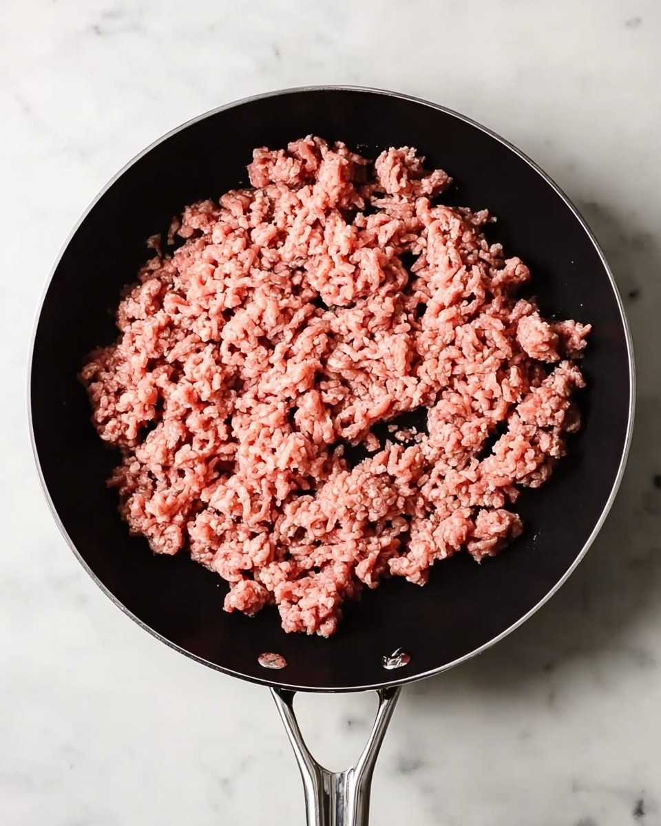 A top view of a black frying pan with two silver handles on a white marbled surface. Inside the pan, there is one layer of raw ground meat spread evenly, showing a mix of pink and light brown colors with a rough, crumbly texture. The pan takes up most of the frame, highlighting the meat's fresh and uneven clumps. photo taken with an iphone --ar 4:5 --v 7