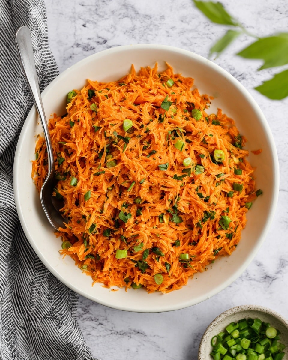 The image shows a white bowl filled with a bright orange shredded carrot salad mixed with small green pieces of herbs and chopped green onions. The carrots and green bits create a textured and colorful mix, with the carrot shreds layered evenly throughout. A silver spoon rests inside the bowl on the left side. The bowl sits on a white marbled surface with a grey striped cloth on the left and a small bowl of chopped green onions on the bottom right. A green leaf is placed at the top right corner. The lighting is natural and soft, making the colors of the salad stand out clearly photo taken with an iphone --ar 4:5 --v 7
