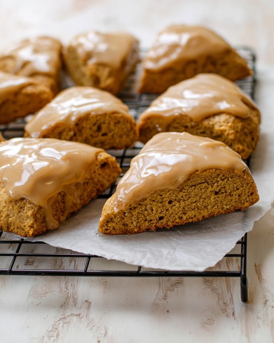 The image shows eight triangular brown scones with a slightly rough texture, each topped with a smooth, shiny light brown glaze that spreads unevenly over the top of the scones. They are arranged in two rows on a black wire cooling rack that is placed over parchment paper, all set on a white marbled surface. The scones look soft and thick, with a dense crumb visible on the cut edges. The focus is on the front row, making the scones in the background appear slightly blurred. photo taken with an iphone --ar 4:5 --v 7