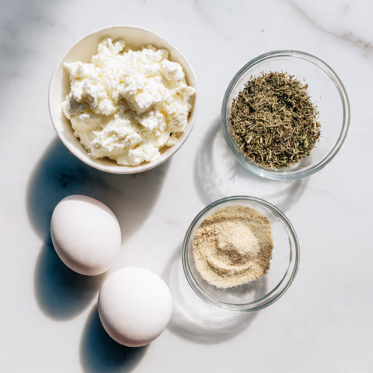 The image shows three simple cooking ingredients arranged on a white marbled surface. In the top left, there is a white container filled with chunky, white cottage cheese. To the right, a clear glass bowl holds three distinct piles of dry spices: light beige garlic powder, dried green herbs, and a pale yellow seasoning. Below these, two white eggs rest close together on the marble surface. The scene is bright and clean, with minimal shadows. photo taken with an iphone --ar 4:5 --v 7