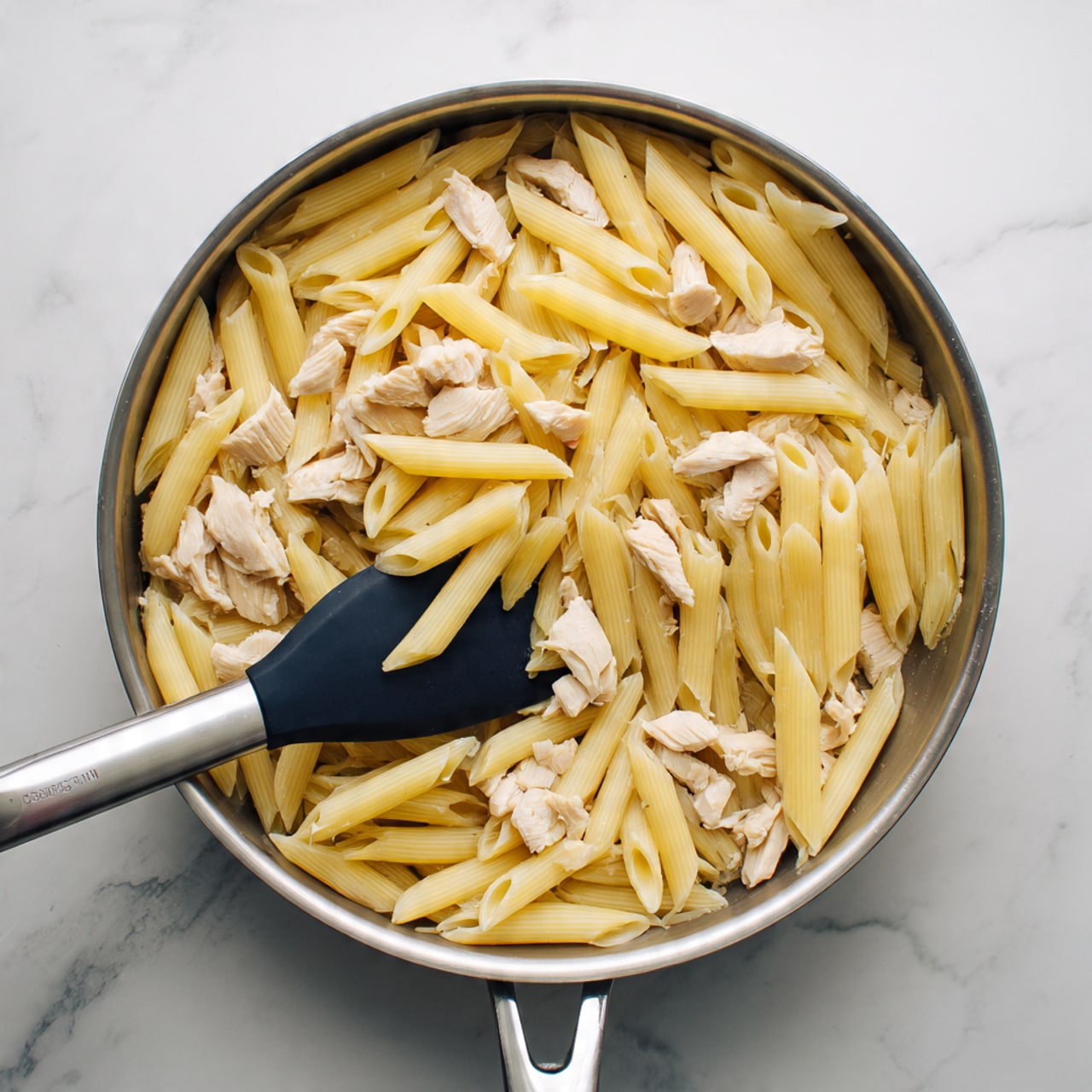 A shiny metal pan sits on a white marbled surface, filled with a mix of light yellow penne pasta and small pieces of white chicken scattered evenly through the dish. A black spatula with a silver handle is resting inside the pan, partly lifting some pasta. The pasta pieces are smooth and slightly glossy, showing a cooked texture, while the chicken pieces are soft and irregular in shape. The pan handle stretches out to the left side of the image, and the lighting highlights the slight shine on the pasta and chicken. photo taken with an iphone --ar 4:5 --v 7