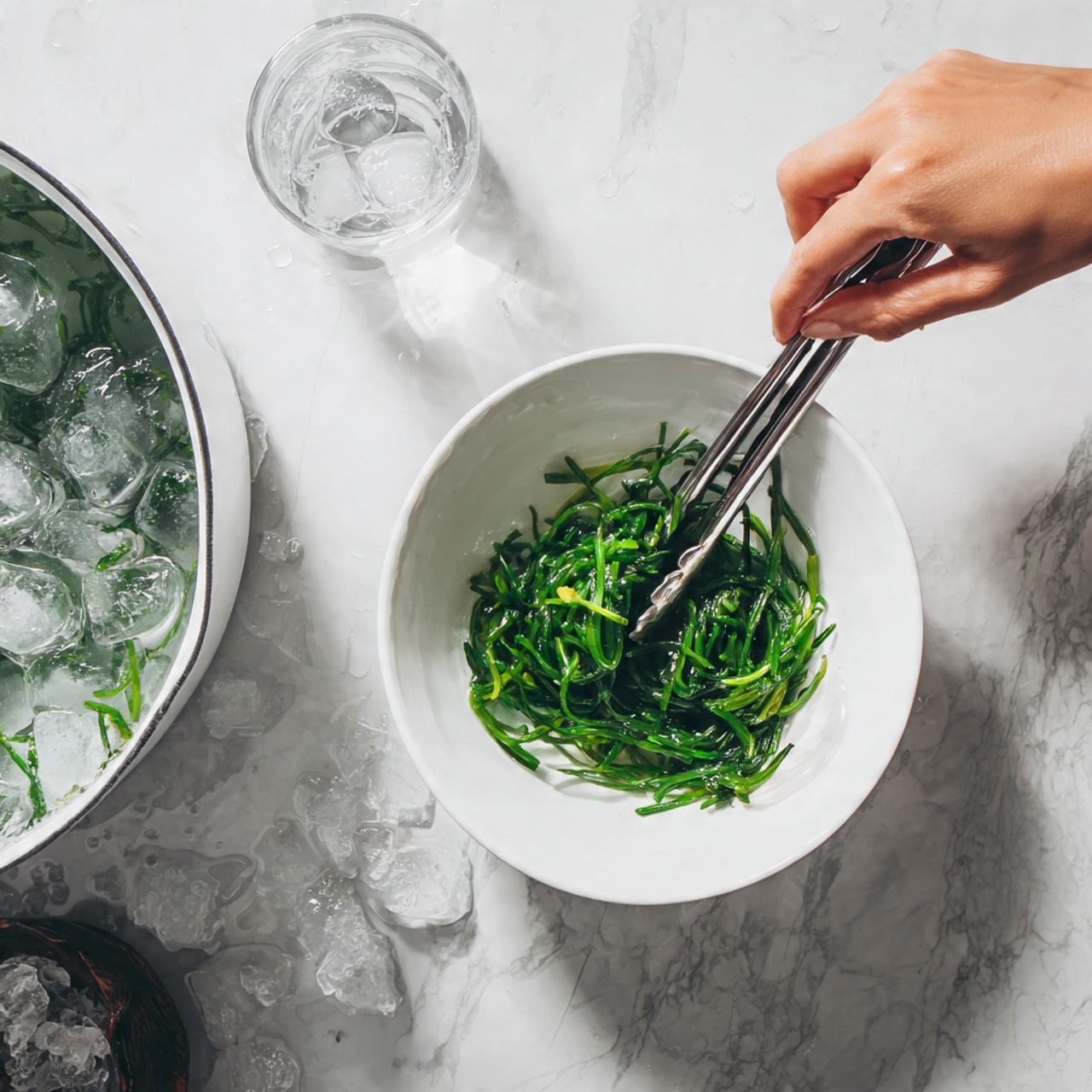 A close-up of a woman's hand holding metal tongs with bright green cooked leafy vegetables above a white bowl filled with clear ice water. To the left, there is a white pot with more cooked green leaves in water, sitting on a white marbled surface. The scene shows the vegetables being placed into the ice water for cooling. photo taken with an iphone --ar 4:5 --v 7