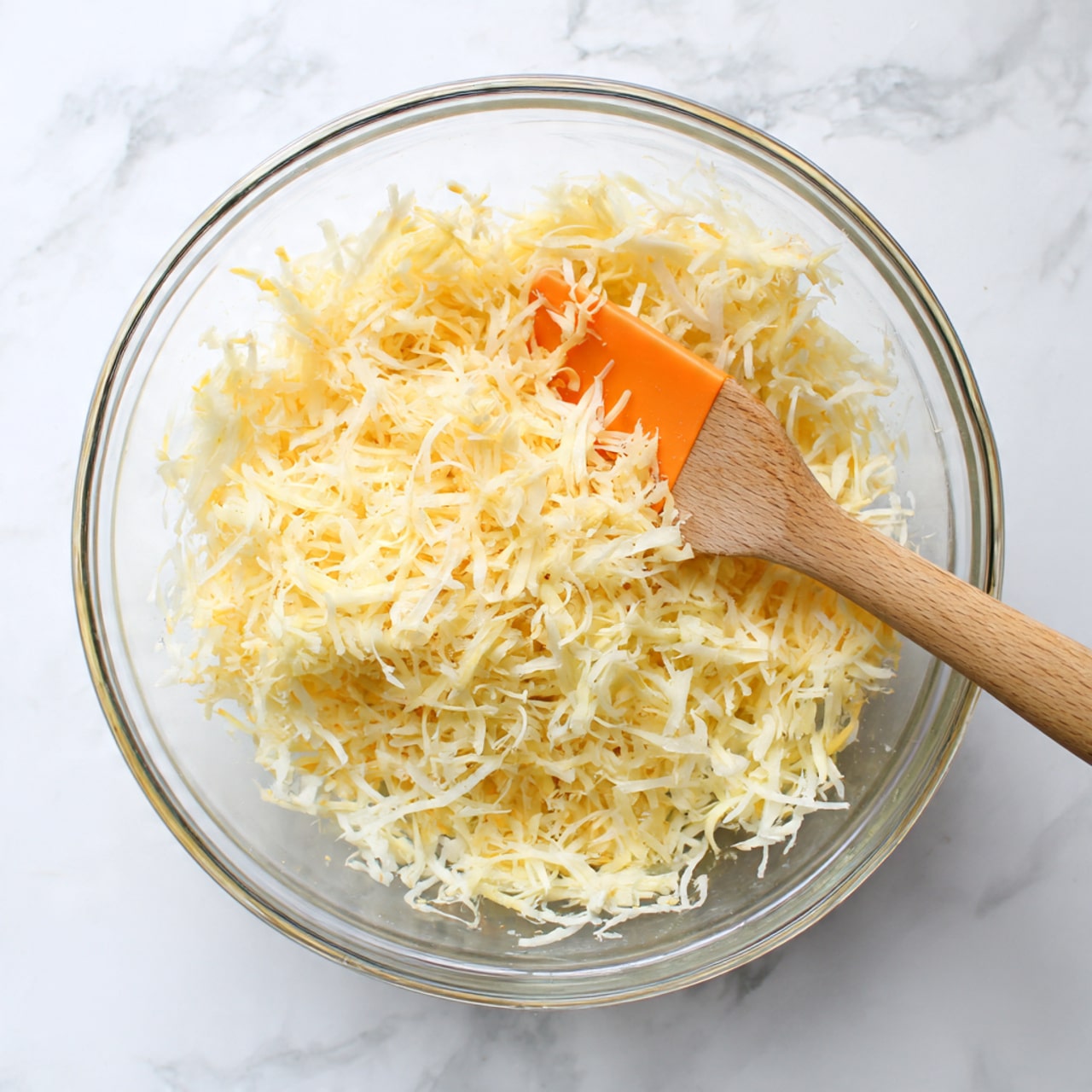 A clear glass bowl filled with a mix of shredded light yellow and pale white ingredients, which look like grated potatoes or cheese. The texture appears moist and stringy, with the strands lightly clumping together. A wooden spatula with an orange silicone head rests inside the bowl, partially buried in the mix, with the handle stretching out to the right. The bowl sits on a white marbled surface. photo taken with an iphone --ar 4:5 --v 7