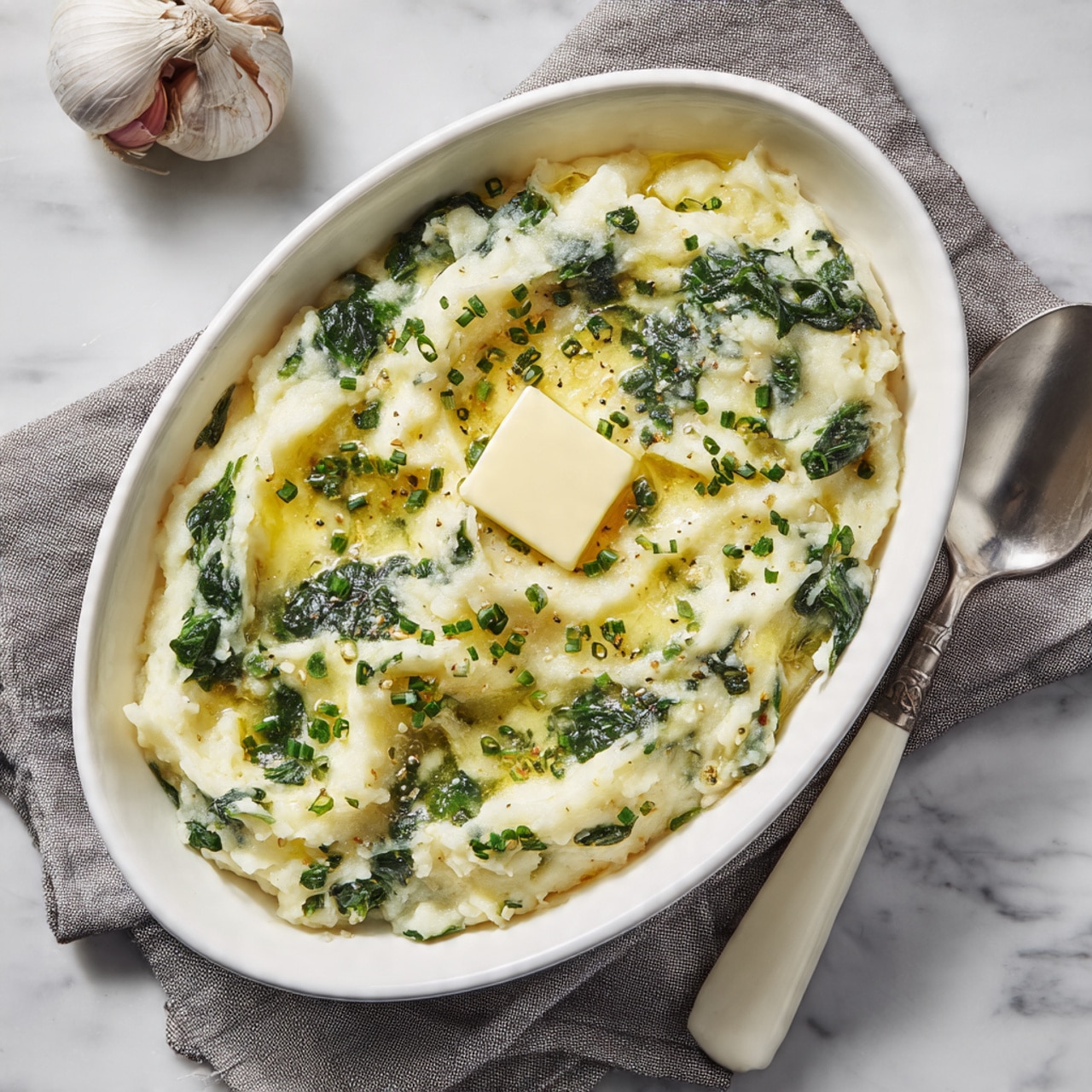 A close-up image of a white bowl full of mashed potatoes mixed with green herbs and leafy vegetables, showing a textured and creamy layer with visible small chunks and bits of greens mixed in evenly. A silver spoon is lifting a portion of the mashed potatoes from the bowl, highlighting the soft and slightly chunky texture with leafy green pieces scattered throughout. The bowl is placed on a white marbled surface, with part of a gray cloth visible on the left side in the background. photo taken with an iphone --ar 4:5 --v 7