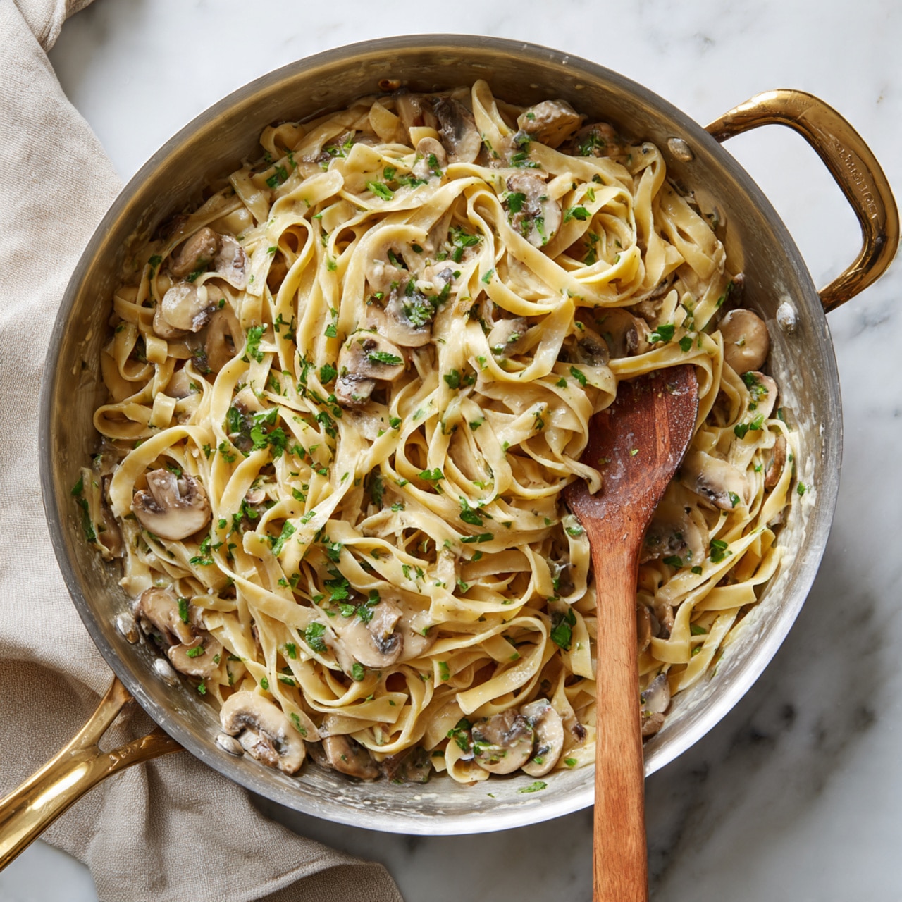 The image shows a shiny silver pan filled with creamy pasta mixed with small pieces of mushrooms and herbs. A woman's hand holds black and silver tongs lifting a bundle of light yellow pasta strands coated in a creamy white sauce with bits of mushrooms and green herbs. The pan sits on a light gray cloth over a white marbled surface, with a clean white background. photo taken with an iphone --ar 4:5 --v 7