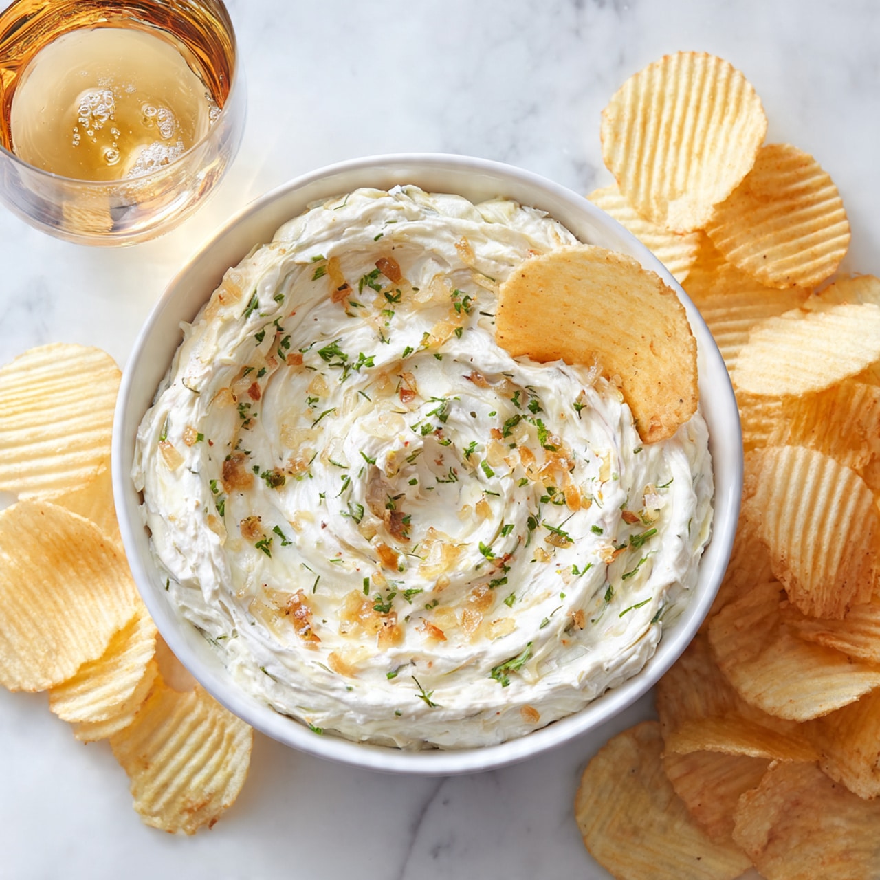 A white bowl filled with creamy white dip that has small bits of green herbs and brown crispy pieces on top, resting on a white marbled surface. A woman's hand is dipping a pale yellow potato chip into the thick, textured dip. Around the bowl, there are some scattered potato chips and a blurred yellow and black chip bag in the background. Photo taken with an iphone --ar 4:5 --v 7