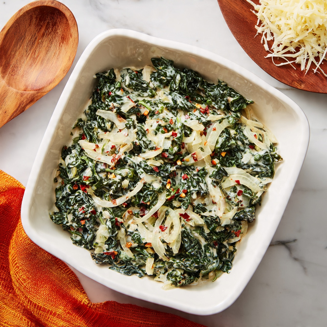 A shiny silver pan filled with cooked dark green kale leaves mixed with a creamy white sauce. Lightly translucent white onion slices are spread throughout the kale, giving extra texture and color contrast. The kale looks soft and well-coated with the sauce, which clings to the leaves and onions evenly. The pan rests on a white marbled surface, adding a clean and fresh look to the image. Photo taken with an iphone --ar 4:5 --v 7