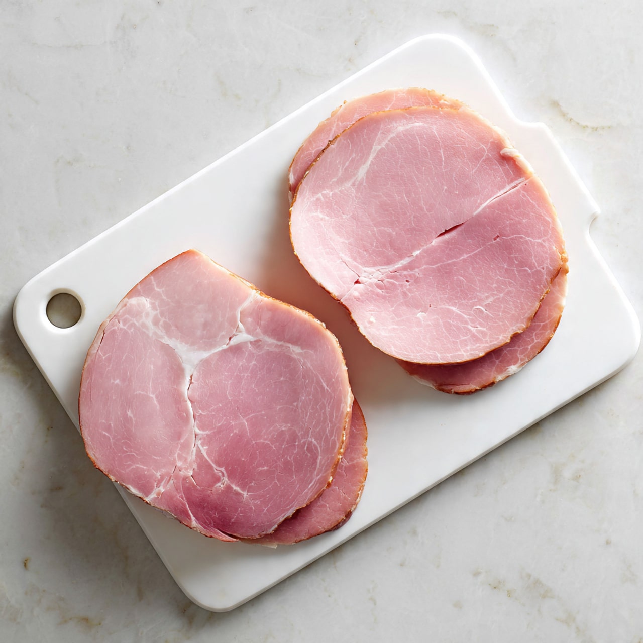 Two large slices of pink ham are placed on a white cutting board with rounded corners and a handle cutout on the left side. Each slice is cut again into two halves, showing smooth, slightly marbled surfaces with light and dark pink areas and small natural holes. The cutting board sits on a white marbled textured surface. The overall look is simple, clean, and fresh, with soft light and natural shadows. photo taken with an iphone --ar 4:5 --v 7