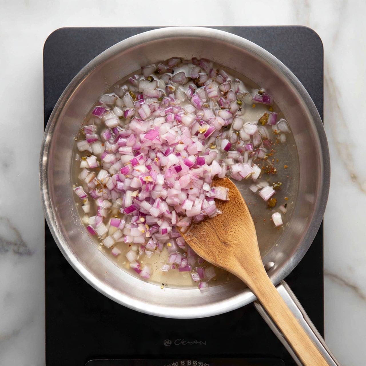 A shiny silver pan sits on a black stove burner, filled with small pieces of red onion and white bits stirring in a translucent liquid. A wooden spoon with a textured surface rests inside the pan, partially mixing the colorful mix, with its handle stretching out over the pan's edge. The background shows a white marbled surface, adding brightness to the scene. photo taken with an iphone --ar 4:5 --v 7
