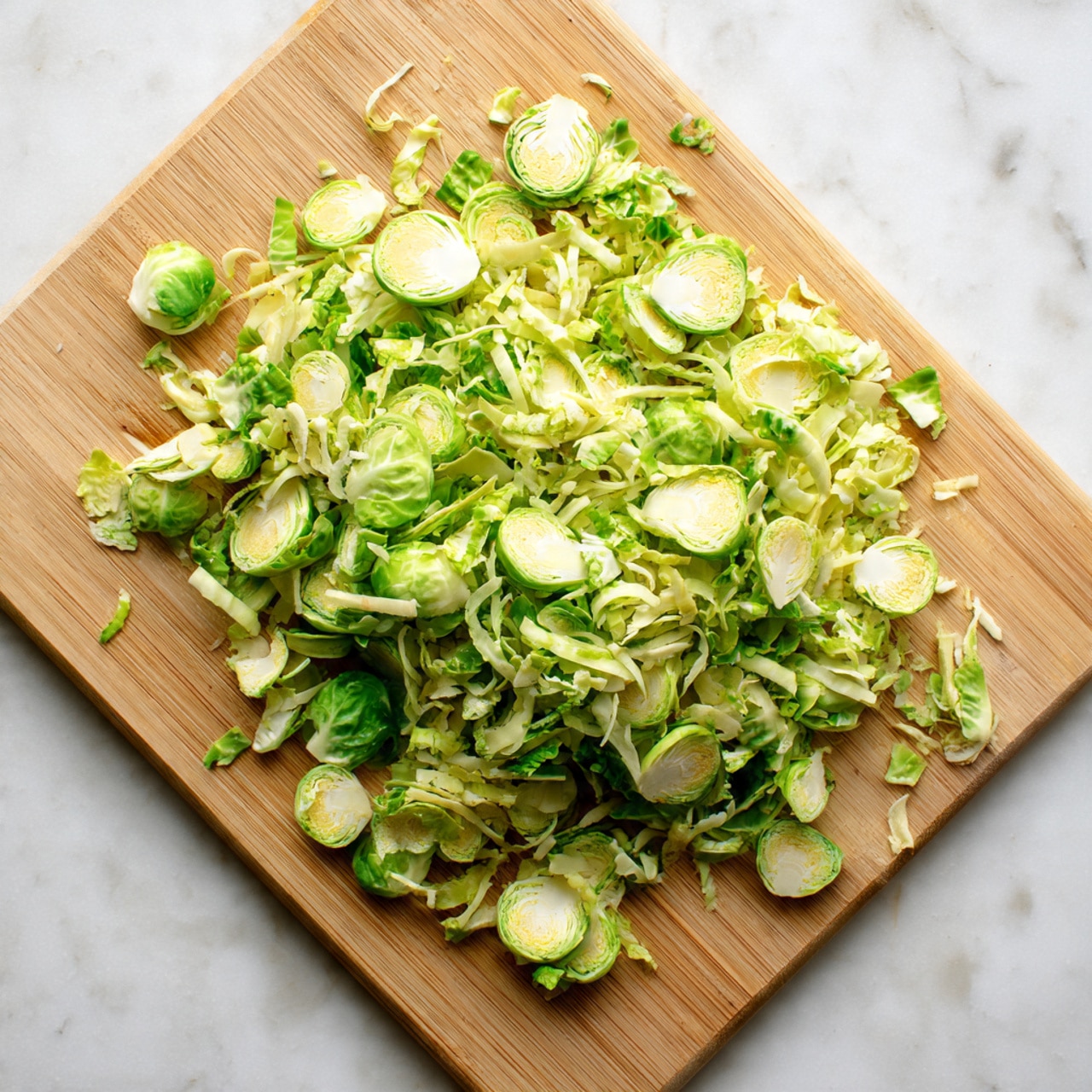 A pile of very thinly sliced pale green Brussels sprouts is spread across a light wooden cutting board, showing a mix of round and curly layers with some white inner parts visible. The cutting board is on a white marbled surface, and the texture of the sprouts looks fresh and crisp with small shreds and slices layered unevenly together, creating a natural, loose mound photo taken with an iphone --ar 4:5 --v 7