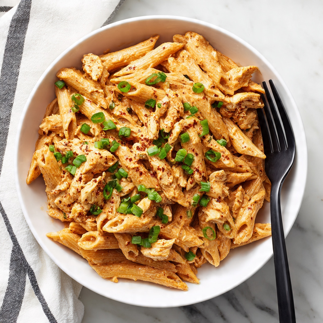A white bowl holds a creamy pasta dish with about three layers. The base layer is short tube-shaped penne pasta, coated in an orange-tan sauce mixed evenly. The second layer includes small pieces of cooked chicken mixed in with the pasta. On top, small bright green chopped herbs, likely green onions, add spots of fresh color. A black fork rests inside the bowl with its handle leaning on the edge. The background is a white marbled surface with a folded white and black striped cloth nearby. Photo taken with an iphone --ar 4:5 --v 7