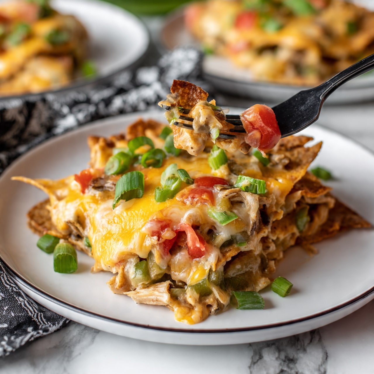 A white plate with a black rim holds a layered serving of melted yellow cheese, chunks of chicken, chopped green bell peppers, and diced tomatoes mixed with creamy sauce. The top layer shows crisp, slightly browned tortilla chips partially covered by cheese and vegetables, with a few fresh green onion pieces sprinkled on top. A black fork is poking into the dish, lifting some of the cheesy mixture. The plate sits on a white marbled surface with a black and white patterned cloth nearby. Another plate with the same dish is partially visible in the corner. Photo taken with an iphone --ar 4:5 --v 7
