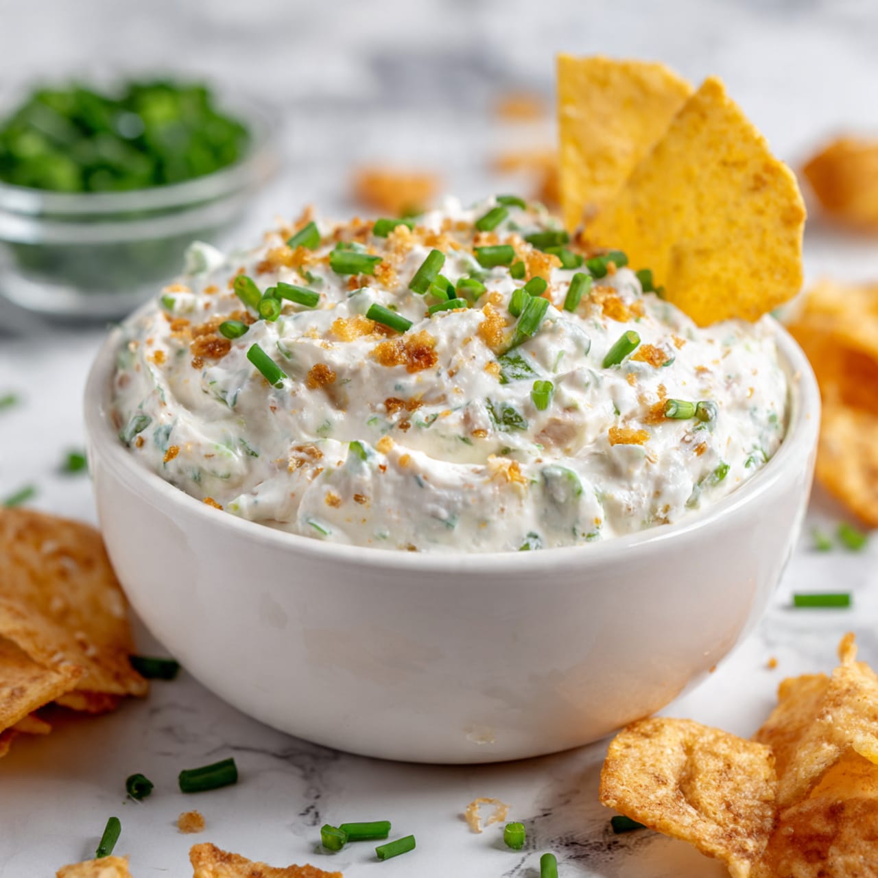 A close-up image of a white bowl filled with a creamy white dip mixed with small golden brown pieces, topped with bright green chopped chives. One yellowish chip is placed partially inside the dip on the right side. The bowl sits on a white marbled texture with some scattered chips and green garnish pieces around it. In the background, there is a clear glass bowl with more green garnish. Photo taken with an iphone --ar 4:5 --v 7