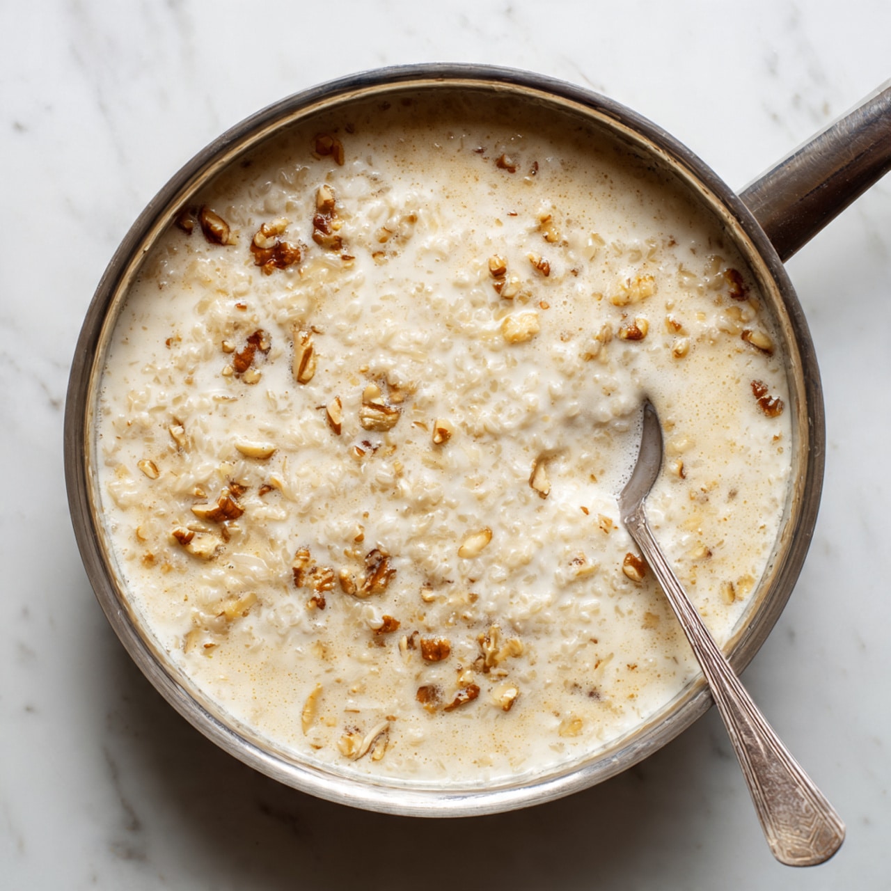 A metal pan filled with creamy beige rice pudding mixed with small pieces of nuts, giving it a lumpy texture. A silver spoon is resting inside the pan on the right side, partially submerged in the pudding. The pan is placed on a white marbled surface, which adds a clean and bright background to the image. The pudding appears thick and rich with small crunchy bits spread all over. photo taken with an iphone --ar 4:5 --v 7
