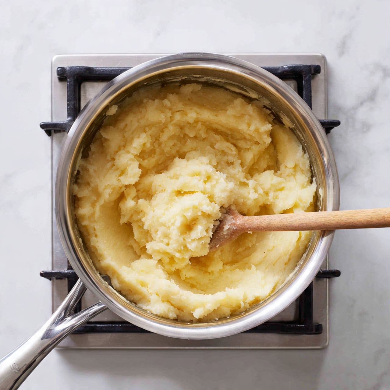 A shiny silver pot sits on a stove with black grates. Inside the pot, there is a thick, pale yellow mashed mixture with a slightly lumpy texture. A wooden spoon with a light brown handle rests inside the pot, partly buried in the mashed mixture. The stove is next to a white marbled countertop. The scene looks like food is being cooked or mixed. photo taken with an iphone --ar 4:5 --v 7